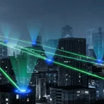 Visualizzazione fotorealistica di una rete WPCN: una stazione base centrale emette onde di energia wireless (verdi brillanti) verso molteplici dispositivi utente (piccoli cerchi blu luminosi) sparsi in un ambiente urbano stilizzato di notte. Profondità di campo, obiettivo 35mm, toni blu e grigi duotone, focus nitido sulle onde energetiche.