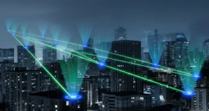 Visualizzazione fotorealistica di una rete WPCN: una stazione base centrale emette onde di energia wireless (verdi brillanti) verso molteplici dispositivi utente (piccoli cerchi blu luminosi) sparsi in un ambiente urbano stilizzato di notte. Profondità di campo, obiettivo 35mm, toni blu e grigi duotone, focus nitido sulle onde energetiche.