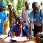 Fotografia concettuale, obiettivo 50mm con profondità di campo, che mostra diverse persone (un operatore sanitario locale, un funzionario governativo, un rappresentante di ONG) attorno a un tavolo in Liberia, le loro espressioni riflettono significati divergenti e tensioni di potere sottostanti, luce naturale filtrata.