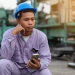 Fotografia realistica di un operaio industriale birmano sulla quarantina, in pausa lavoro all'esterno di una fabbrica a Mandalay. Guarda pensieroso il suo vecchio cellulare che tiene in mano, forse dopo aver letto un SMS. Luce del tardo pomeriggio, obiettivo prime 50mm, profondità di campo ridotta per sfocare lo sfondo industriale. Atmosfera di riflessione e speranza.