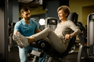 Fotografia realistica di una donna anziana sorridente (circa 70 anni) che esegue un esercizio di potenziamento del quadricipite su una macchina in palestra, assistita da un fisioterapista. Luce calda e motivante. Obiettivo prime 35mm, profondità di campo che isola leggermente i soggetti, colori naturali.