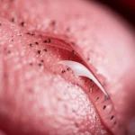 Macro fotografia (obiettivo macro 100mm) di una goccia di gel trasparente e viscoso, contenente minuscole particelle sospese (rappresentanti il complesso farmaco-ciclodestrina), che aderisce a una superficie rosa e leggermente texturizzata che simula la mucosa della gola. L'illuminazione è controllata e morbida per enfatizzare la trasparenza e l'adesione, alta definizione, messa a fuoco selettiva sulla goccia.