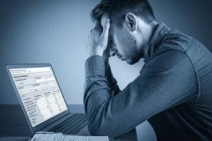 Ritratto fotografico di un giovane (20-25 anni) dall'aspetto pensieroso e leggermente sopraffatto, seduto a una scrivania con un laptop aperto su un modulo online dall'aspetto burocratico. Obiettivo 35mm, effetto profondità di campo, bicromia blu e grigio per trasmettere formalità e malinconia.