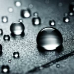 Immagine macro fotorealistica di una superficie metallica high-tech con rivestimento in lega Cu-Sn. Gocce d'acqua sferiche perfette rotolano via, dimostrando proprietà superidrofobiche e autopulenti. La superficie mostra una micro-nano texture complessa. Illuminazione drammatica che evidenzia la lucentezza metallica e le gocce. Obiettivo macro 105mm, alta definizione.