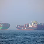 Fotografia teleobiettivo di due moderne navi portacontainer che navigano vicine in una rotta di potenziale collisione in una giornata nebbiosa nello Stretto di Gibilterra, tracciamento del movimento, velocità dell'otturatore elevata per congelare l'azione, luce drammatica e diffusa dalla nebbia.
