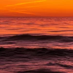 Immagine grandangolare della superficie dell'oceano al tramonto, con onde dolci che riflettono colori caldi arancioni e rossi, simbolo del riscaldamento globale degli oceani, lunga esposizione per acqua liscia come seta, obiettivo 20mm, messa a fuoco nitida sull'orizzonte lontano.