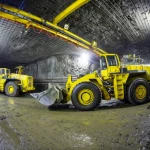 Panoramica fotorealistica dell'interno di una miniera metallifera sotterranea moderna e meccanizzata. Si vedono macchinari pesanti come un LHD (Load Haul Dumper) in azione e sistemi di supporto del soffitto. Obiettivo grandangolare 15mm, illuminazione artificiale potente che rivela la vastità e la complessità dell'ambiente, messa a fuoco nitida su tutta la scena.
