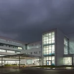 Immagine simbolo della resilienza ospedaliera: una struttura ospedaliera moderna e illuminata che resiste a una tempesta metaforica (nuvole scure, pioggia), con personale medico visibile ma calmo all'interno. Fotografia architettonica, obiettivo grandangolare 18mm, lunga esposizione per nuvole mosse, focus nitido sull'ospedale.