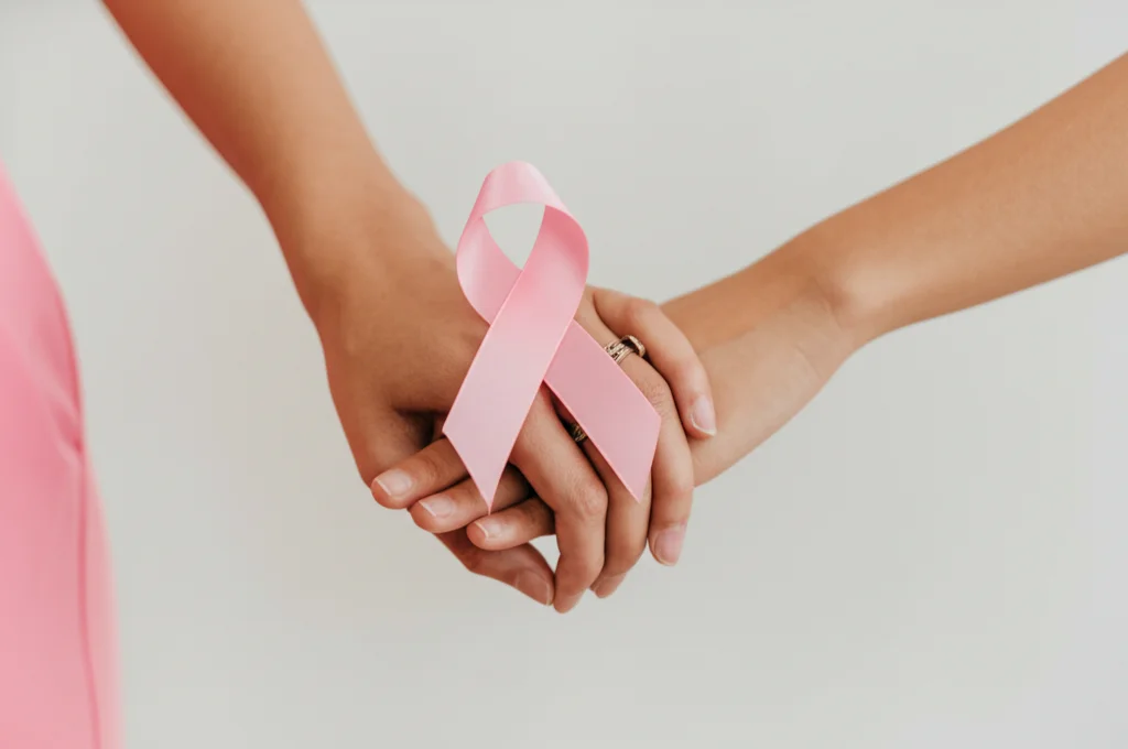 Immagine simbolica: due mani femminili diverse, una curata con anelli (alto SES) e una più semplice (basso SES), che stringono insieme un nastro rosa simbolo della lotta al cancro al seno. Sfondo neutro e morbido, obiettivo prime 50mm, profondità di campo accentuata, illuminazione diffusa che crea un'atmosfera di riflessione e speranza.
