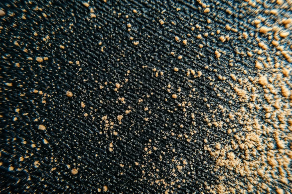 Scatto macro con lente 85mm, che mostra una lastra di gomma NBR nera texturizzata parzialmente ricoperta da fine polvere nano di pula di riso (nRH) di colore marrone chiaro, alto dettaglio, messa a fuoco precisa, illuminazione controllata da studio che enfatizza il contrasto tra i due materiali innovativi.