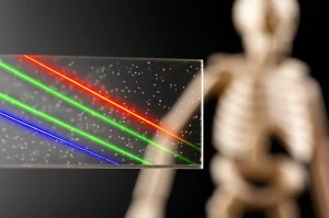 Immagine fotorealistica con obiettivo macro 80mm, alto dettaglio e illuminazione controllata, che mostra un campione di vetro borosilicato trasparente con nanoparticelle di ZnO disperse, che devia simbolicamente delle linee luminose rappresentanti radiazioni gamma. Sullo sfondo, una sfocata struttura ossea stilizzata suggerisce l'applicazione biomedica. Profondità di campo ridotta per focalizzare sul vetro.