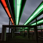Vista grandangolare fotorealistica (obiettivo 15mm) di una complessa rete luminosa di tubi interconnessi (gas o acqua) che si estende in lontananza sotto un cielo tempestoso e incerto, a simboleggiare la domanda fluttuante. Messa a fuoco nitida, effetto di lunga esposizione sulla 'luminescenza' all'interno dei tubi.