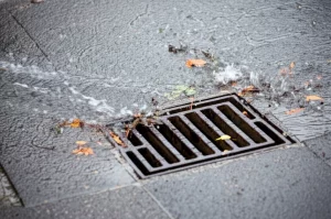 Acqua piovana che scorre lungo un marciapiede urbano grigio verso un tombino metallico, trascinando piccole foglie e detriti, ripresa dall'alto con obiettivo grandangolare 24mm, messa a fuoco nitida sull'acqua in movimento, luce naturale diffusa dopo un acquazzone.
