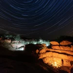 Fotografia ad ampio raggio, 24mm, del riparo roccioso di Itramuka a Singida, Tanzania, al tramonto, con la silhouette di archeologi che lavorano meticolosamente in uno scavo illuminato da luci artificiali, lunga esposizione per catturare le stelle e il colore del cielo.