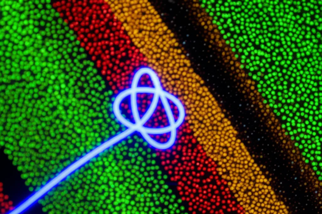 Rappresentazione artistica e scientifica di un complesso nodo luminoso tridimensionale (simile a un nodo a trifoglio) che emerge da un pattern di luce granulare (speckle) caotico e fluttuante sullo sfondo. Il nodo è formato da una linea luminosa continua che rappresenta le singolarità di coerenza. Obiettivo prime, 35mm, profondità di campo ridotta per focalizzare sul nodo, illuminazione drammatica con contrasto elevato, stile quasi film noir ma a colori vivaci per il nodo.