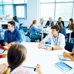 Aula universitaria moderna e luminosa con diversi piccoli gruppi di studenti di medicina (circa 4-5 per gruppo) seduti attorno a tavoli rotondi, impegnati attivamente in una discussione collaborativa durante una sessione di Team Based Learning. Alcuni indicano schermi di tablet, altri scrivono su fogli. Un docente si muove tra i gruppi osservando e interagendo. Fotografia grandangolare 24mm, messa a fuoco nitida su tutta la scena, luce naturale abbondante, atmosfera dinamica e positiva di apprendimento attivo.