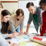 Immagine fotorealistica: Un gruppo eterogeneo di giovani adulti collabora attorno a un tavolo pieno di schizzi, appunti e prototipi. Le loro espressioni sono concentrate (metacognizione), le posture impegnate (motivazione), le interazioni sicure (autoefficacia), simboleggiando l'intersezione di questi fattori nel valorizzare la creatività in azione. Obiettivo prime (35mm), profondità di campo ridotta, luce naturale che illumina la scena.