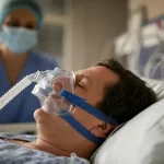 Primo piano di una maschera CPAP trasparente indossata da un paziente sdraiato in un letto d'ospedale, si intravede un operatore sanitario con mascherina sullo sfondo sfocato, luce clinica ma calda, obiettivo 50mm, profondità di campo ridotta per focalizzare sulla maschera.