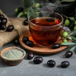 Composizione still life con polvere di noccioli d'oliva in una ciotola di ceramica, noccioli interi sparsi, e una tazza di tè di nocciolo d'oliva fumante su sfondo neutro e rustico, lente macro 100mm, alta definizione, messa a fuoco precisa sui dettagli della polvere e del liquido, illuminazione laterale morbida che crea ombre delicate.