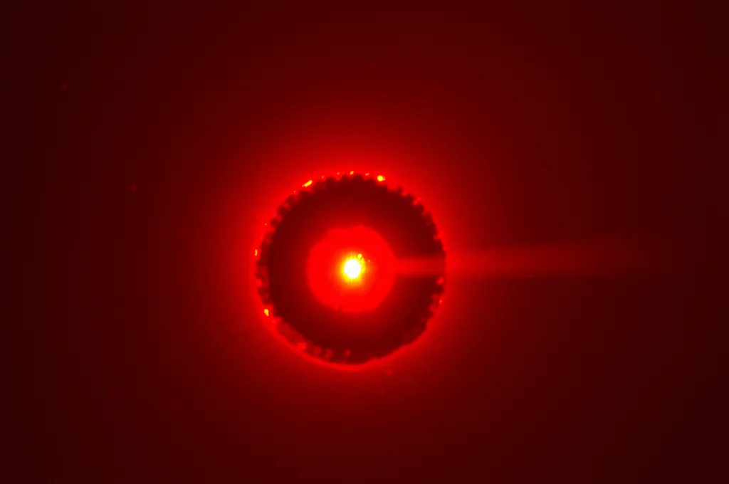 Immagine fotorealistica, teleobiettivo zoom 200mm, che cattura il debole bagliore rosso della luce NIR focalizzata su un modello di tumore al fegato di topo durante una sessione di terapia fototermica in un laboratorio di ricerca. Tracciamento del movimento del fascio di luce, profondità di campo ridotta, ambiente sterile e scientifico.