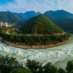 Immagine panoramica del fiume Duliu in Cina che scorre attraverso un paesaggio montuoso, con una sezione trasformata in un lago artificiale da una grande diga idroelettrica visibile, contrasto tra acqua calma a monte e fiume naturale a valle, obiettivo grandangolare 20mm, luce del tardo pomeriggio, alta definizione, messa a fuoco nitida.