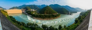 Immagine panoramica del fiume Duliu in Cina che scorre attraverso un paesaggio montuoso, con una sezione trasformata in un lago artificiale da una grande diga idroelettrica visibile, contrasto tra acqua calma a monte e fiume naturale a valle, obiettivo grandangolare 20mm, luce del tardo pomeriggio, alta definizione, messa a fuoco nitida.
