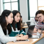 Studenti EFL cinesi in un istituto professionale superiore impegnati in un'attività di apprendimento misto basato su SPOC. Un mix di interazione online su dispositivi (laptop, tablet) e discussione offline in un'aula moderna e luminosa. Fotografia di persone, obiettivo zoom 35mm, profondità di campo media per mostrare sia gli studenti in primo piano che l'ambiente, luce naturale che entra dalle finestre, espressioni concentrate e collaborative.