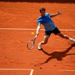 Azione dinamica di un giocatore di tennis che colpisce la palla con un rovescio potente su un campo in terra battuta, catturata con teleobiettivo zoom 200mm, fast shutter speed per congelare il movimento della palla e della racchetta, tracciamento dell'azione, luce solare pomeridiana intensa che crea ombre nette.