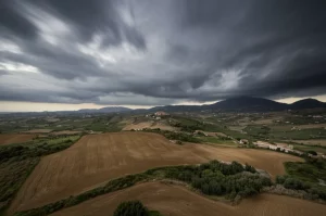 Veduta aerea grandangolare di un paesaggio siciliano sotto un cielo tempestoso scuro, con nuvole cariche di pioggia che incombono su colline e campi, obiettivo grandangolare 15mm, lunga esposizione per nuvole mosse, messa a fuoco nitida sul paesaggio, luce drammatica del tardo pomeriggio.