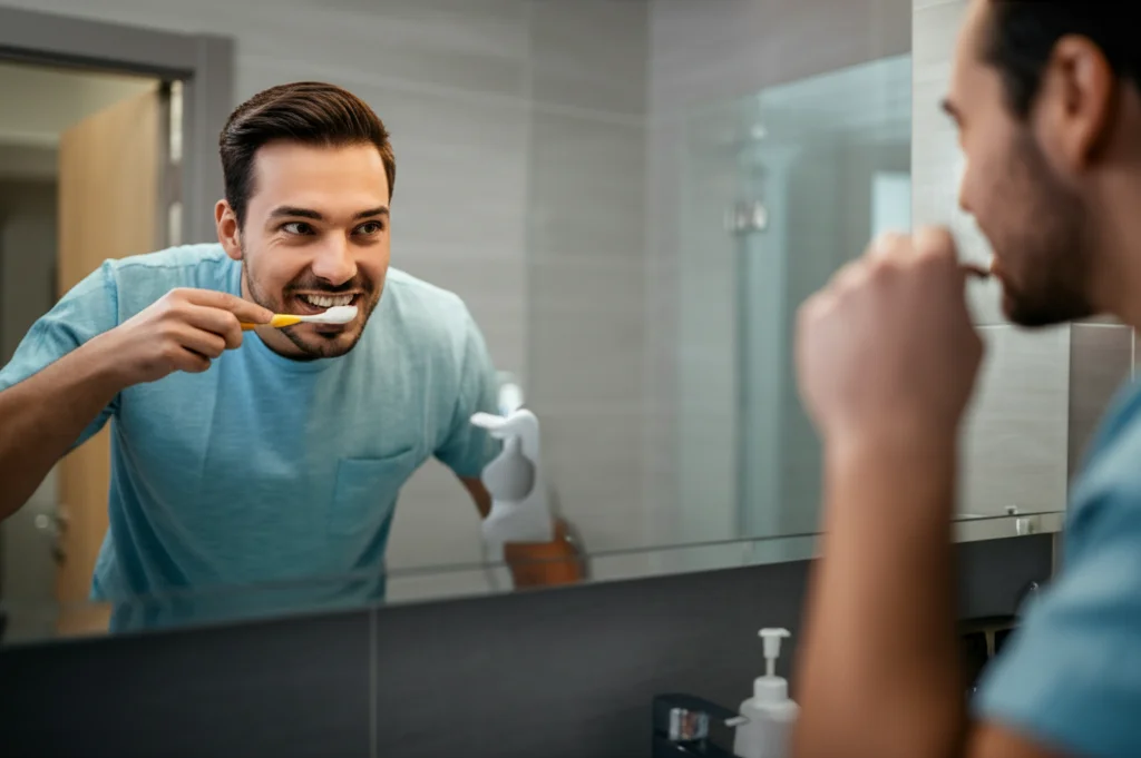 Una persona adulta sorridente che si lava i denti davanti allo specchio del bagno. Luce naturale del mattino. Prime lens, 35mm, depth of field, focus sul viso e sullo spazzolino.