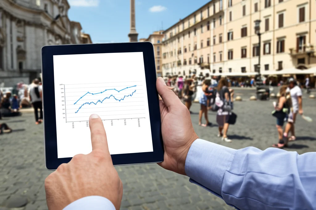 Un decisore politico o un analista osserva attentamente un grafico di previsione del turismo (linea che prosegue nel futuro) su un tablet, con lo sfondo leggermente sfocato di una famosa piazza italiana (es. Piazza Navona) piena di turisti. Ritratto, obiettivo zoom 50mm, profondità di campo che isola il soggetto e il tablet, colori naturali e luce diurna.