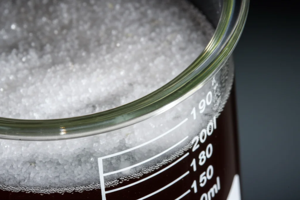 Fotografia still life di cristalli bianchi purissimi di cloruro di ammonio (NH4Cl) raccolti dopo il processo di elettroriduzione dei nitrati in acido, macro lens 100mm, illuminazione controllata per evidenziare la purezza, accanto a un becher contenente la soluzione elettrolitica.