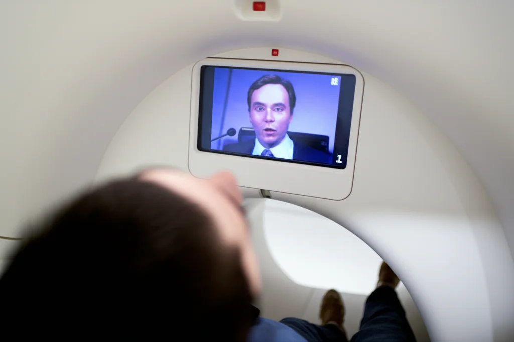 Immagine fotorealistica di una persona all'interno di uno scanner fMRI, vista leggermente dall'alto, con uno schermo visibile che proietta il volto di uno speaker durante un discorso. Illuminazione controllata tipica di un laboratorio medico/di ricerca, focus nitido sullo schermo e sul riflesso negli occhi del partecipante. Obiettivo prime 35mm, profondità di campo ridotta per isolare il soggetto.