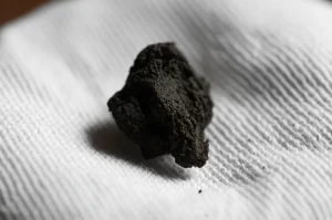 Immagine fotorealistica, lente macro 100 mm, dettagli elevati, messa a fuoco precisa, illuminazione controllata, che mostra una piccola quantità di "argilla benedetta" scura e umida da Boho tenute attentamente in un pezzo di semplice stoffa bianca, che suggerisce sia alla tradizione che al potenziale scientifico.