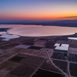 Veduta aerea paesaggistica del Bacino di Yanqi, Xinjiang, al tramonto. Obiettivo grandangolare 18mm, esposizione lunga per catturare le sfumature arancioni e viola del cielo e ammorbidire la superficie del lago Bosten in lontananza. Messa a fuoco nitida sull'orizzonte, mostrando il mosaico geometrico dei campi agricoli irrigati che contrasta con le zone desertiche circostanti, simboleggiando la cruciale gestione delle risorse idriche in un ambiente arido. Luce calda e dorata del sole calante.