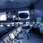 Immagine fotorealistica di una sala controllo missione spaziale moderna, schermi multipli mostrano dati orbitali complessi della Luna e della sonda KPLO (Danuri). Tecnici concentrati al lavoro. Illuminazione controllata, stile cinematografico, obiettivo 35mm per un'ampia vista della sala, profondità di campo che sfoca leggermente lo sfondo.