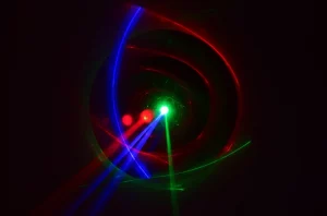 Rappresentazione artistica e fotorealistica di tre fasci di luce coerente (rosso, verde, blu) che convergono e interagiscono intensamente in un punto focale su uno sfondo scuro, creando pattern di interferenza complessi e luminosi. Obiettivo prime 50mm, profondità di campo ridotta per focalizzare l'interazione, illuminazione drammatica per evidenziare la non linearità.