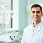 Ritratto di un giovane studente di odontoiatria sorridente in camice bianco, in un moderno studio dentistico universitario luminoso. Obiettivo prime 35mm, profondità di campo per sfocare leggermente lo sfondo, luce naturale che entra da una finestra.