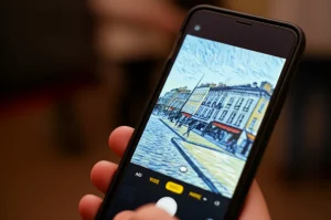 Smartphone che mostra un'app di style transfer in azione, trasformando una foto normale di un paesaggio urbano in un dipinto nello stile di Van Gogh. Primo piano sullo schermo luminoso, sfondo leggermente sfocato. Illuminazione controllata, obiettivo prime 35mm, profondità di campo ridotta.