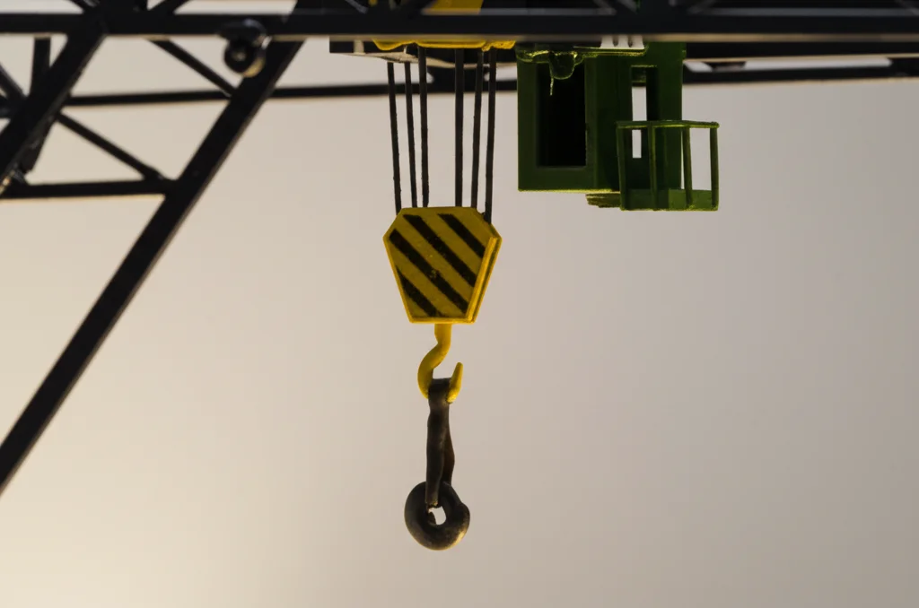Fotografia macro ad alta definizione di un modello in scala di una gru a ponte con un carico a pendolo, illuminazione controllata da studio che evidenzia i dettagli meccanici del carrello e del cavo, obiettivo macro 90mm, messa a fuoco precisa sul carico sospeso.