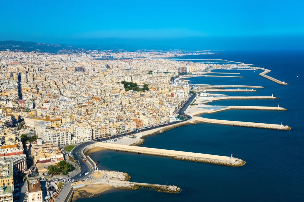 Veduta aerea grandangolare di una città costiera densamente popolata con evidenti difese marittime (muri o dighe). Alcune aree sembrano vulnerabili all'innalzamento del livello del mare. Obiettivo grandangolare 18mm, luce diurna brillante, messa a fuoco nitida sull'interfaccia tra città e mare.