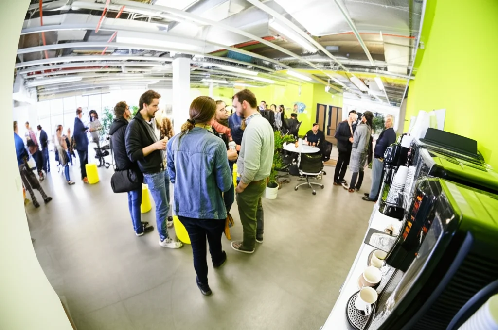 Fotografia grandangolare di un moderno ufficio open space durante una pausa caffè. Diverse persone interagiscono in piccoli gruppi in modo rilassato e amichevole vicino a una macchinetta del caffè. Obiettivo grandangolare 20mm, luce naturale brillante, messa a fuoco nitida sull'ambiente generale per catturare l'atmosfera collaborativa.