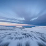 Fotografia paesaggistica grandangolare che cattura un solo sciano di fondo che si addestrasse vigorosamente all'alba attraverso un vasto e incontaminato altopiano nevoso nelle montagne norvegesi. Lente grandangolare, lunghezza focale di 18 mm, focus acuto in tutta la scena, effetto a lungo esposizione che leviga le nuvole di alba colorate. Luce mattutina drammatica e bassa creazione di lunghe ombre. Trasmette la solitudine, l'immenso sforzo, la dedizione e la scala epica dell'allenamento di resistenza in natura.