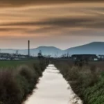 Fotografia panoramica, wide-angle 10mm, che cattura il contrasto tra un'area industriale moderna e prospera (simbolo del Nord Italia) e una zona rurale o meno sviluppata con infrastrutture datate (simbolo del Sud Italia), separate metaforicamente da una linea divisoria naturale come un fiume o una catena montuosa. Luce drammatica del tramonto, cielo suggestivo con nuvole mosse (long exposure), sharp focus su entrambi i lati del paesaggio.