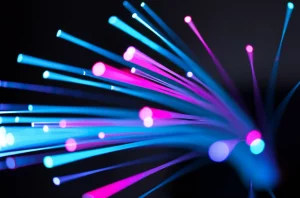 Visualizzazione artistica di impulsi luminosi che viaggiano ad altissima velocità attraverso i multipli core di una fibra ottica. Effetto motion blur per indicare velocità, colori vivaci (ciano, magenta) per rappresentare i dati, sfondo scuro high-tech. Teleobiettivo zoom 150mm, fast shutter speed per congelare parzialmente il movimento ma con scie luminose.