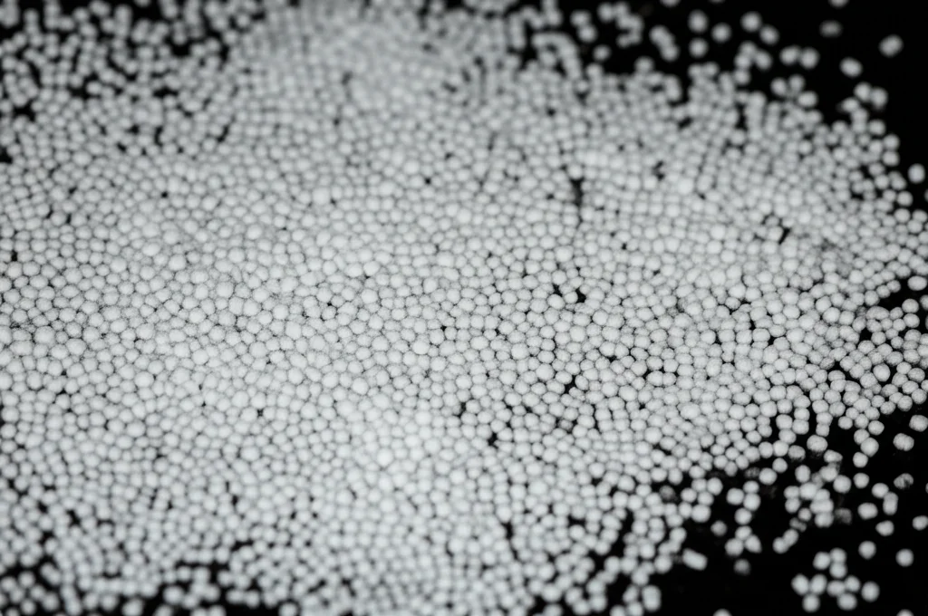 Immagine macro di particelle di PMMA riciclato, alcune fini e polverose, altre più grandi e irregolari, disposte su una superficie da laboratorio. Illuminazione controllata laterale per evidenziare la texture, obiettivo macro 60mm, alta definizione.