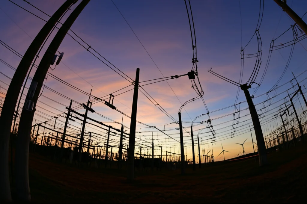 Veduta grandangolare di una moderna smart grid al tramonto, con linee elettriche illuminate, trasformatori in silhouette e turbine eoliche in lontananza sotto un cielo colorato, obiettivo grandangolare 15mm, lunga esposizione per scie luminose, cielo con nuvole sfumate e vibranti.