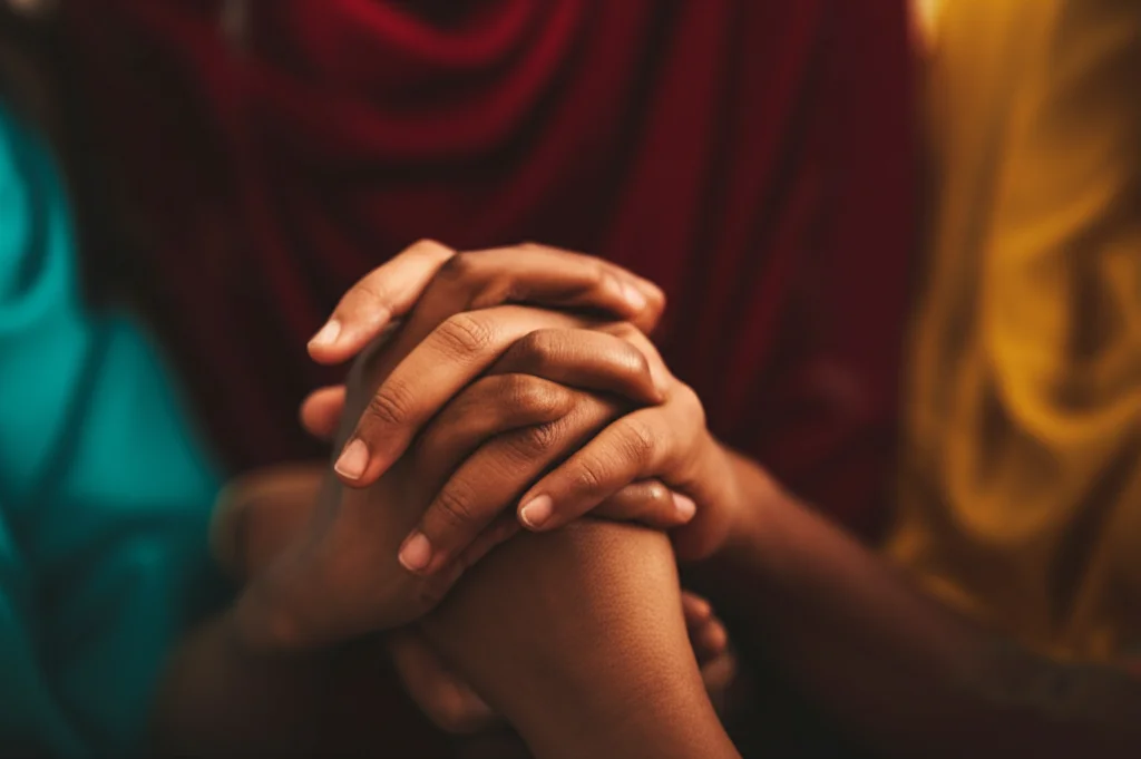 Primo piano sulle mani intrecciate di due donne etiopi durante una sessione di gruppo di supporto, simboleggiando connessione e solidarietà. Macro lens 60mm, messa a fuoco precisa sui dettagli delle mani, illuminazione calda e controllata per enfatizzare il legame.