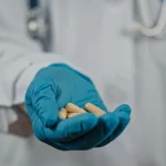 Primo piano di capsule dell'antivirale Molnupiravir tenute in una mano guantata da un professionista sanitario in un ambiente clinico. Luce morbida, obiettivo prime 50mm, profondità di campo che sfoca leggermente lo sfondo per concentrarsi sul farmaco.