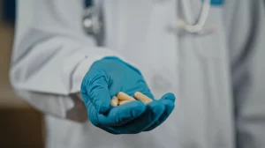 Primo piano di capsule dell'antivirale Molnupiravir tenute in una mano guantata da un professionista sanitario in un ambiente clinico. Luce morbida, obiettivo prime 50mm, profondità di campo che sfoca leggermente lo sfondo per concentrarsi sul farmaco.